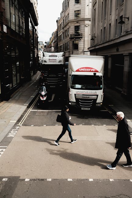 View from a street-level perspective showing a narrow urban street with tall, light-colored buildings on either side. In the foreground, two pedestrians are crossing at a pedestrian crossing; one is a woman carrying a shoulder bag, and the other is an older man with white hair. In the background, there is a white van with the logo and website of Uniserve, partially blocking the width of the street, positioned behind a black motorcycle parked on the road. Behind the van, a larger truck with the Becton Transport logo is visible, suggesting vehicle parking or loading for a home relocation or removals service. The scene is lit by daylight, capturing a typical city environment suited for furniture transport and packing during a house move, supported by the presence of professional vehicles associated with Marylebone Removals.