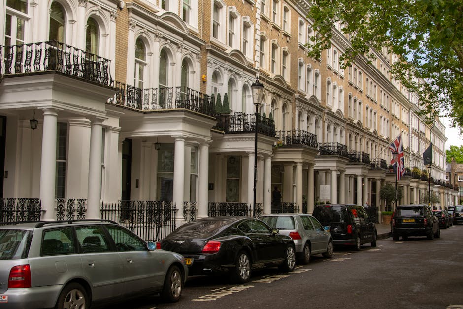 Exterior view of a row of elegant Georgian-style townhouses in Marylebone, London, with white facades, black wrought-iron balconies, and large windows. The street shows parked cars lined along the pavement, including a silver estate, a black coupe, and other vehicles. A black van is partially visible near the sidewalk, suggesting furniture transport or loading associated with home relocation services. The scene is well-lit with natural daylight, and a street lamp stands next to a flagpole flying the Union Jack, indicating a typical British urban residential area. The presence of the parked vehicles and the organized street scene imply the environment for house removals or furniture transport tasks, which a professional removals company like Marylebone Removals might facilitate.