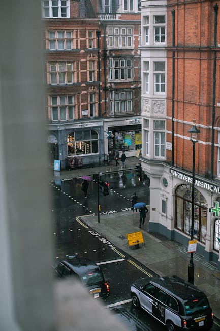 View of a rainy street scene in Marylebone, London, captured from an upstairs window. The street features a row of red-brick and white-facade buildings, with ground-floor commercial spaces including a pharmacy with a green cross sign. Pedestrians with umbrellas walk along the pavement, and a black car is parked on the road. The wet pavement reflects the surrounding buildings and streetlights, indicating recent rain. Visible are street signs and a temporary yellow barrier on the curb, alongside a loading zone, which is relevant for home relocation and furniture transport logistics. The scene exemplifies the urban environment where Marylebone Removals offers house removal and packing services, supporting efficient moving processes in busy city locations.