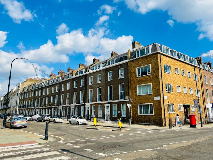 Exterior view of a row of elegant Georgian-style townhouses in Marylebone, London, with white facades, black wrought-iron balconies, and large windows. The street shows parked cars lined along the pavement, including a silver estate, a black coupe, and other vehicles. A black van is partially visible near the sidewalk, suggesting furniture transport or loading associated with home relocation services. The scene is well-lit with natural daylight, and a street lamp stands next to a flagpole flying the Union Jack, indicating a typical British urban residential area. The presence of the parked vehicles and the organized street scene imply the environment for house removals or furniture transport tasks, which a professional removals company like Marylebone Removals might facilitate.