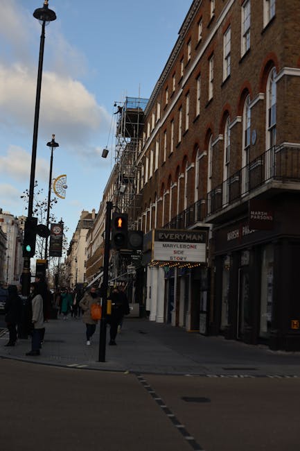 A busy street scene in Marylebone showing a row of multi-storey brick buildings with shopfronts on the ground level, including a sign for Marylebone Storage. The street is populated with pedestrians, some carrying bags, walking on the pavement near a traffic light displaying a red pedestrian signal. There are several street lamps along the sidewalk, and traffic signals are visible at the corner. In the background, scaffolding and construction material can be seen on one of the building facades. The scene is captured during daylight with a partly cloudy sky overhead. This setting illustrates the typical urban environment in Marylebone, with potential for home relocation activities such as furniture transport or packing and moving processes, supported by local removals services like Marylebone Removals.