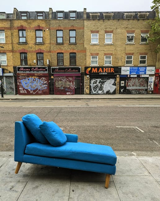 A blue upholstered chaise lounge with two matching cushions positioned on the sidewalk outside a commercial building in Marylebone, London. The furniture is placed directly on the pavement, facing the street with its backrest and extended footrest visible. Behind it, the building features a brick façade with several windows, some of which have white blinds, and multiple closed shopfronts with graffiti on their metal shutters. The scene is set during daylight with natural light illuminating the area, illustrating a typical urban environment. This image relates to home relocation or furniture transport as part of a house removal process, and the furniture appears ready for either disposal or collection during packing and moving activities, with Marylebone Removals potentially involved in the logistics.