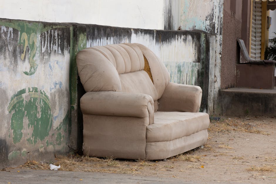 A beige upholstered armchair with visible wear and tear, including torn fabric and stained cushions, is placed outdoors against a weathered graffiti-covered wall with faded green paint and peeling plaster. The chair is situated on a dirt ground with scattered dry leaves and small debris, adjacent to a concrete step leading into a building with a partially visible open doorway. Surrounding the scene are elements of urban decay, such as cracks in the pavement and chipped paint on the wall, reflecting a setting where furniture might be discarded or prepared for removal. The lighting appears natural, providing a clear view of the damaged furniture and rough environment that could be part of a home relocation or furniture disposal process, which the professional team at Marylebone Removals is capable of managing in compliance with local laws regarding furniture disposal in Marylebone and W1.