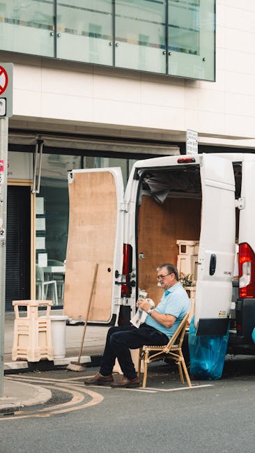 A man sits on a wicker chair beside a white cargo van parked outside a modern building with large glass windows. The van's side door is open, revealing wooden furniture, cardboard boxes, and packing m