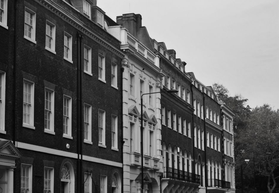 Black and white photograph of a row of historic terraced houses on Marylebone's narrow streets, featuring multi-story brick facades with large sash windows, some with decorative mouldings and small balconies. The street scene shows a street lamp and a few trees in the background, with the pavement in the foreground. This setting depicts an urban environment suitable for home relocation or furniture transport services. The image captures a quiet, residential atmosphere consistent with the kind of property where professional removals, such as those offered by Marylebone Removals, would facilitate the packing, loading, and transportation process of household items, including fragile furniture and appliances, during a house move.