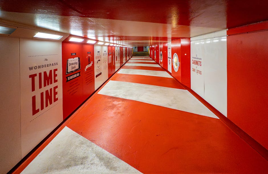 An underground pedestrian tunnel with red and white painted walls and ceiling, featuring various posters and advertisements on the side panels, including one reading 'The Wonderpass Time Line' and another with 'Dinosaur Book'. The floor is patterned with alternating red and white sections, guiding the way through the tunnel. Overhead, the lighting is bright, creating a well-lit environment. The space appears wide enough for moving furniture or boxes, with a clear path extending into the distance. This setting exemplifies indoor logistical spaces used during home relocation or furniture transport processes, with visible signs of environmental preparation for moving activities, and the tunnel's design facilitating smooth passage for搬家 teams and equipment, typical of a city-centre or transport hub environment.