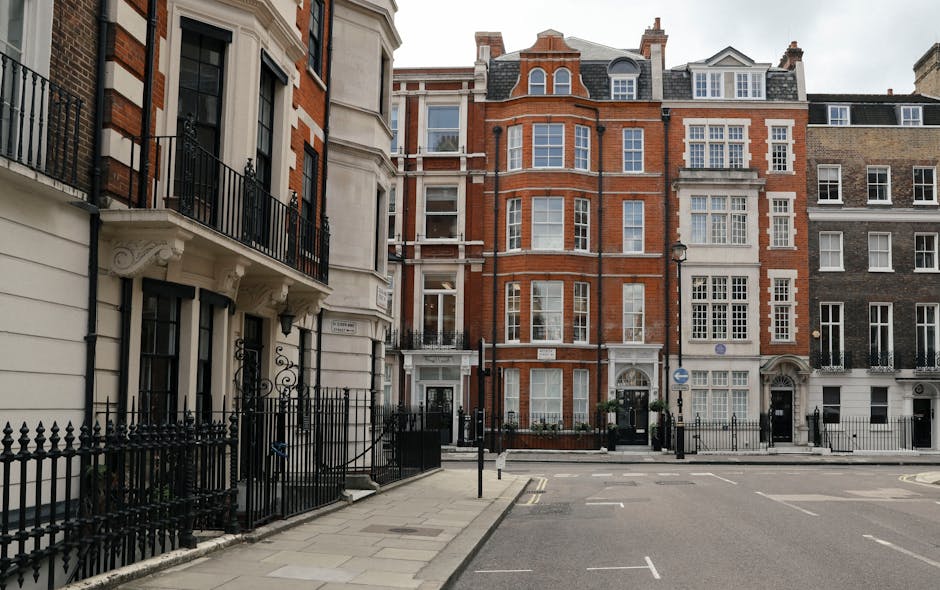 Black and white photograph of a row of historic terraced houses on Marylebone's narrow streets, featuring multi-story brick facades with large sash windows, some with decorative mouldings and small balconies. The street scene shows a street lamp and a few trees in the background, with the pavement in the foreground. This setting depicts an urban environment suitable for home relocation or furniture transport services. The image captures a quiet, residential atmosphere consistent with the kind of property where professional removals, such as those offered by Marylebone Removals, would facilitate the packing, loading, and transportation process of household items, including fragile furniture and appliances, during a house move.