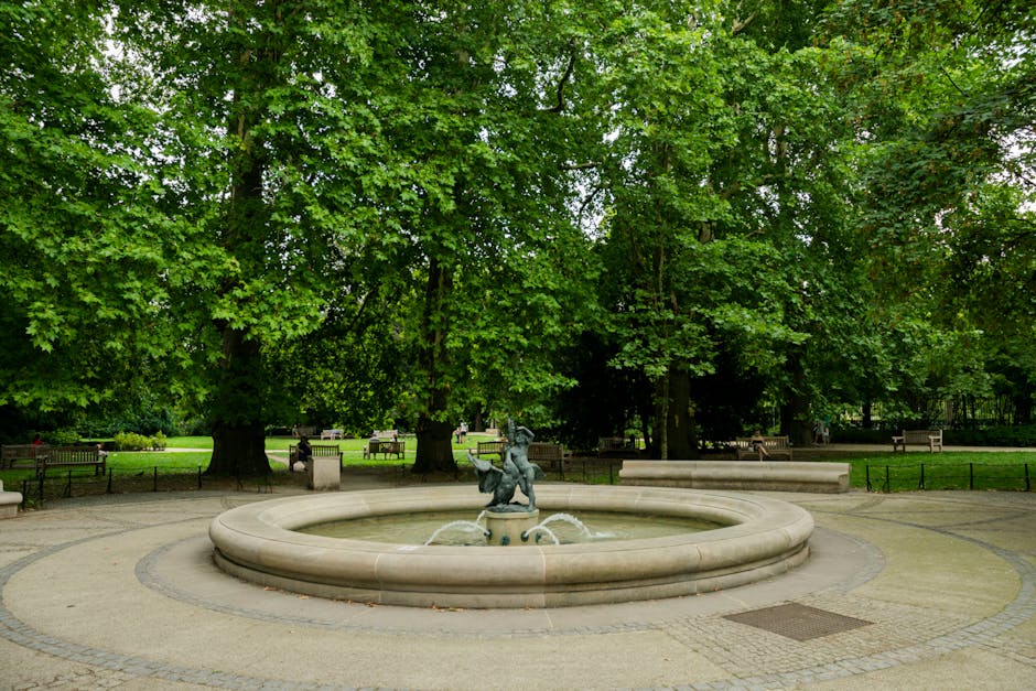 A circular stone fountain with a bronze sculpture of a child and a dog in the centre, surrounded by water spouting from small nozzles, is situated in a park setting. The fountain is positioned on a paved area with cobblestone and concrete surfaces, with several wooden benches and stone planters nearby. Behind the fountain, there are large, leafy green trees providing shade, with a grassy area extending into the distance. The scene appears to be in daylight with natural light filtering through the dense foliage. This image captures a quiet park space that may be used during a house relocation or moving process, where furniture and belongings are temporarily stored or moved through outdoor areas, reflecting the typical environment in Marylebone as seen in the 'Portman Square to Regent's Park' guide by Marylebone Removals.