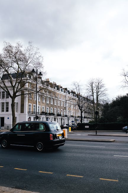 A black London taxi cab parked on a street adjacent to a pavement, with multi-storey residential buildings featuring white window frames and brick facades in the background. Several large, leafless trees are situated along the sidewalk, indicating a late autumn or winter setting. The street has yellow dashed lines and a bollard near the curb. In the foreground, a portion of a building entrance with a white facade is visible, suggesting the proximity to a residential or commercial property. This scene illustrates the typical urban environment where professional removals, such as those offered by Marylebone Removals, may load or unload furniture and packing materials during a home relocation process.