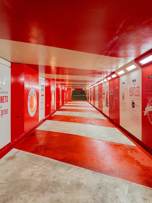 A brightly lit underground passageway with red walls and a matching red ceiling, featuring illuminated rectangular light fixtures along the top edges. The floor is divided into wide alternating red and white stripes. On one side of the corridor, there are various informational posters and signs, some with circular designs, attached to the walls. At the far end, a staircase with dark steps leads upwards, suggesting the exit point. The scene appears to be part of a property that may be involved in home relocation or packing and moving processes, aligning with professional removals services like those offered by Marylebone Removals.