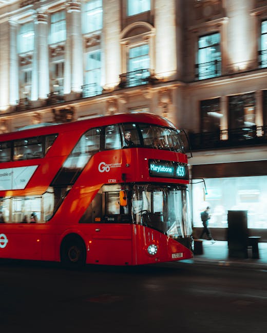 A red double-decker bus with the destination display reading 'Marylebone' is moving along a city street at night. The bus is a typical London black cab style, featuring large windows and the iconic roundel logo on its side. Behind the bus, elegant historic buildings with detailed facades and large windows are illuminated by street lighting, creating a warm glow. Pedestrians are visible walking on the sidewalk, and some are near the bus stop. The scene captures a busy urban environment suitable for home relocation and furniture transport services, highlighting the vibrant city atmosphere where Marylebone Removals offers same-day, urgent move solutions. The lighting and motion blur reflect a bustling, efficient moving process in central London, emphasizing the importance of organized packing and transportation during busy hours.