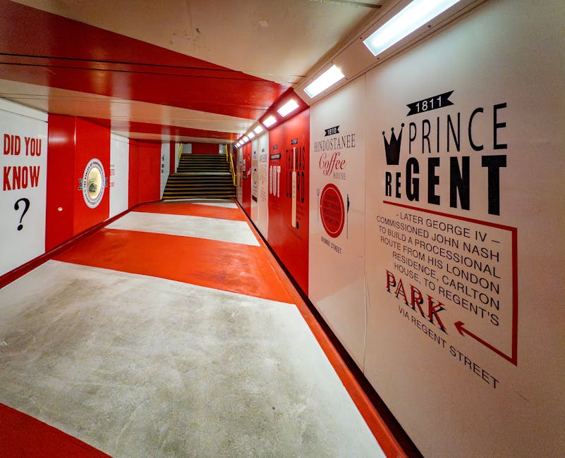 An indoor underground car park featuring a long corridor with red and white painted walls and ceiling. On the right side, large vintage-style signs provide historical information and directions, including a prominent sign with a crown graphic indicating Prince Regent, and an arrow pointing towards the parking area with street name Regent Street. The floor is a combination of smooth concrete and red-painted sections, leading to a staircase at the far end. The ceiling is fitted with fluorescent lighting, illuminating the space evenly. The corridor appears clean and well-maintained, with no furniture or moving equipment visible, creating a neutral environment suitable for loading or unloading items during a home relocation. This setting reflects an urban parking or service area associated with house removals, supporting the logistics of furniture transport and packing and moving processes, as handled by Marylebone Removals.